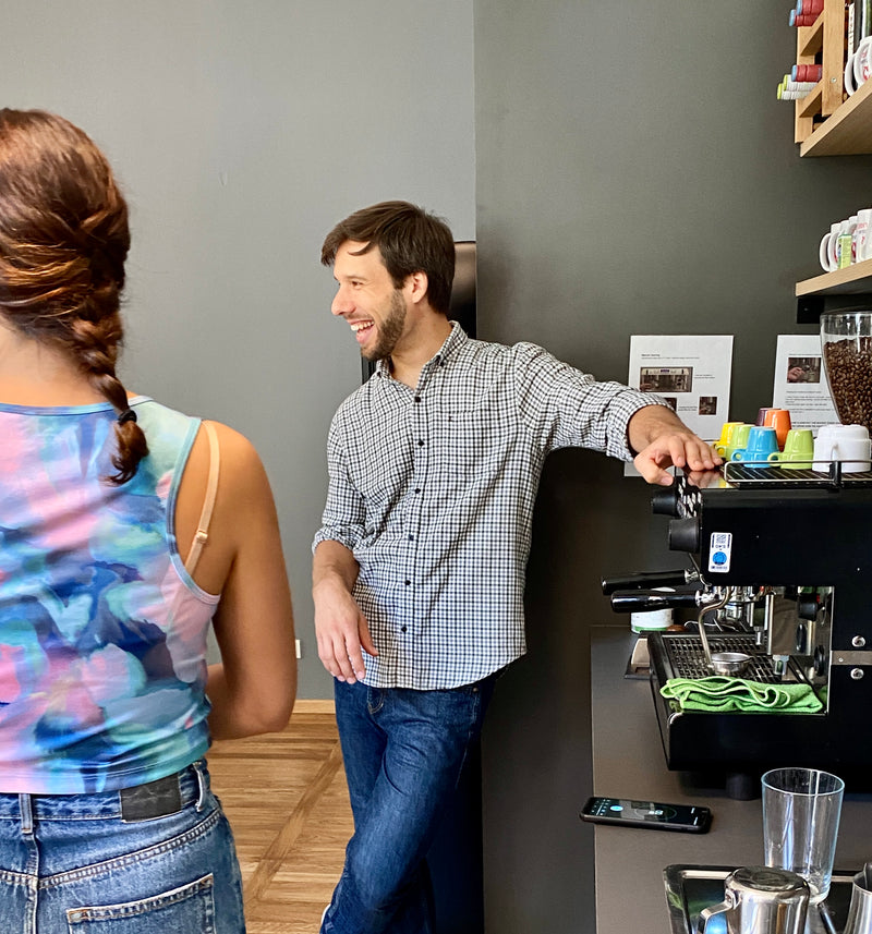 Ein Mann (Sebastian) lehnt entspannt und lachend neben einer professionellen Espresso-Siebträgermaschine und führt ein persönliches Beratungsgespräch mit einer Kundin. Das Bild visualisiert die Rolle von Sortengold als unabhängiger Kaffeepartner, der durch fachlichen Austausch ohne Dogma und eine markenunabhängige Beratung für eine verlässliche Infrastruktur im Büro sorgt.